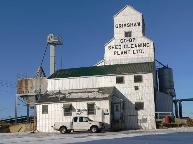 Exterior of Grimshaw Co-op Seed Cleaning Plant. There is a tall, white wooden building with a white pickup truck parked in front of it.